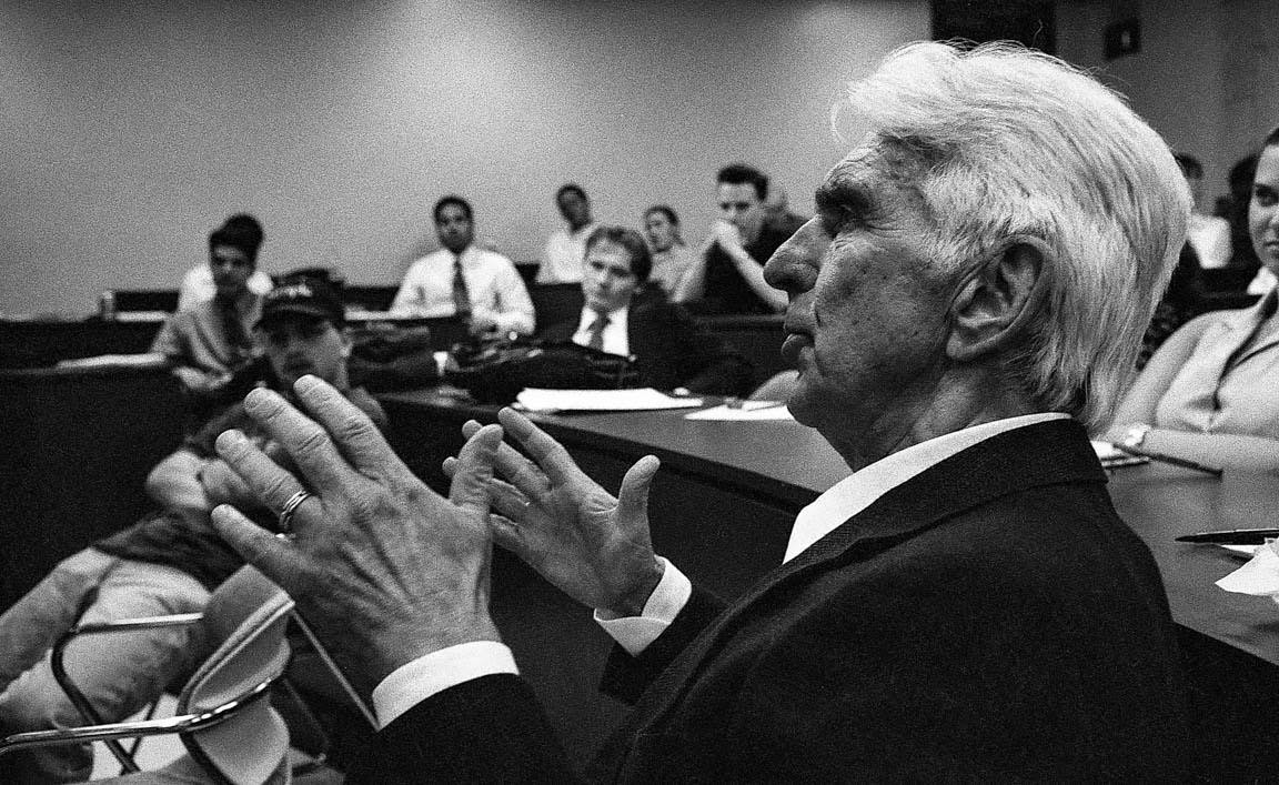 A man in a suit and tie speaks as other people look on.