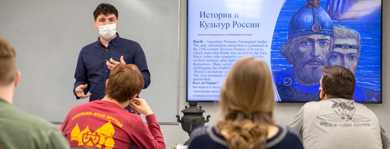 Russian language instructor Randall Rowe at the front of the classroom. Screen with Russian language writing behind him.