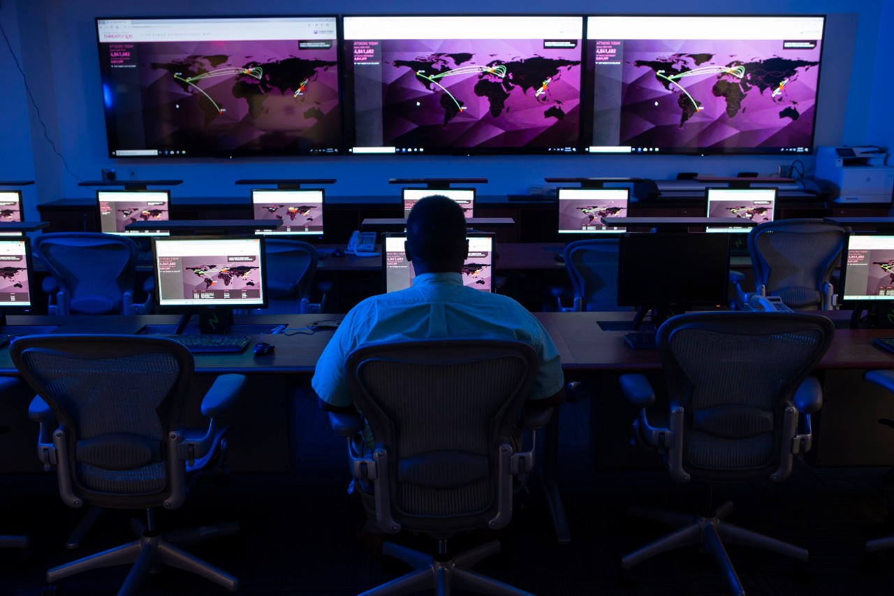 A person sits at a bank of computers with bigger screens on the wall.