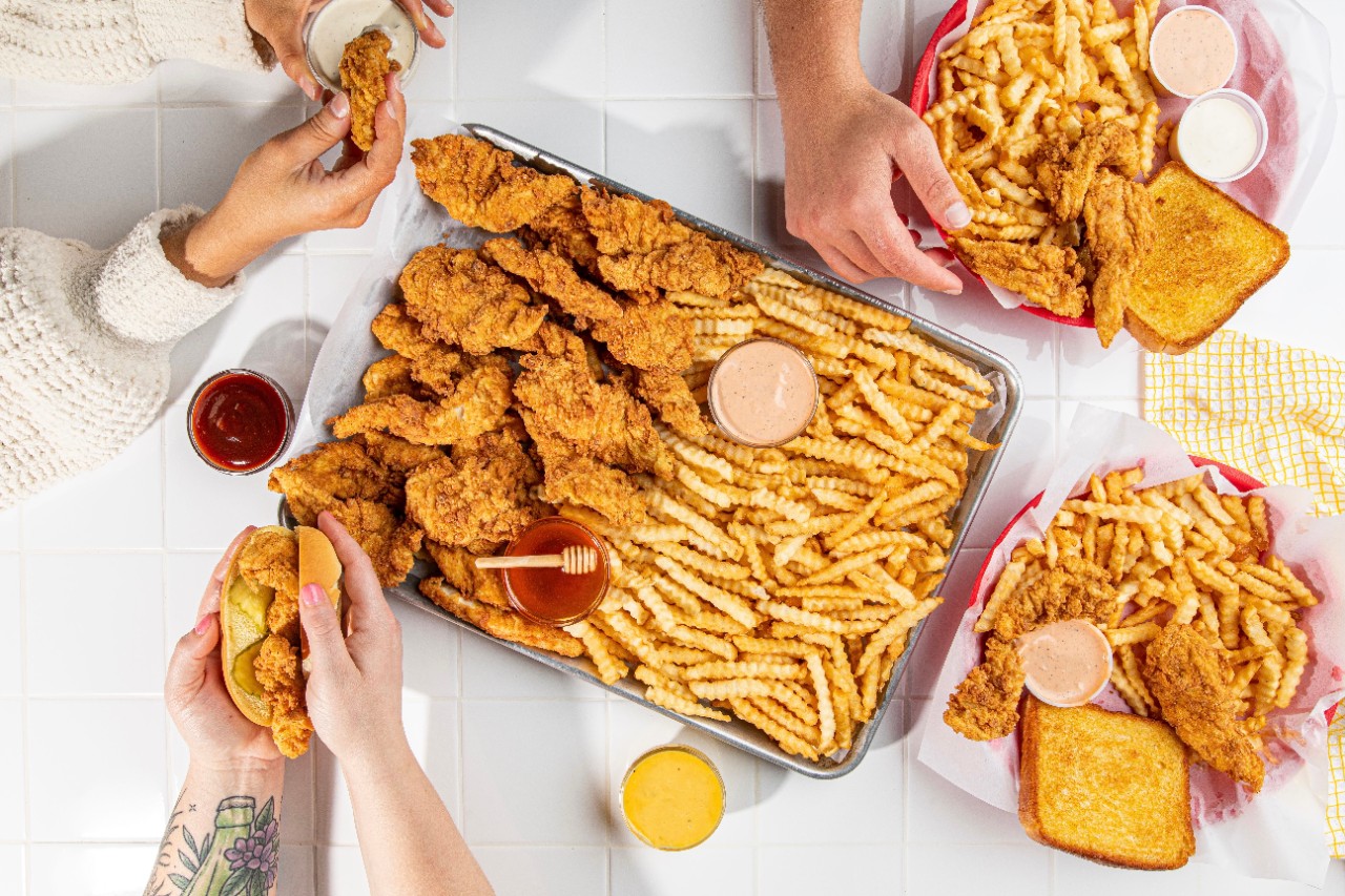 Three sets of human hands eat chicken fingers and fries with sauces.