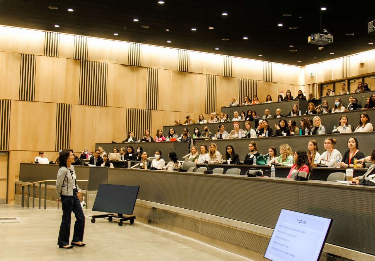A woman with chin length dark hair wearing a white and black sweater and black pants stands in front of a classroom auditorium speaking while students are seated