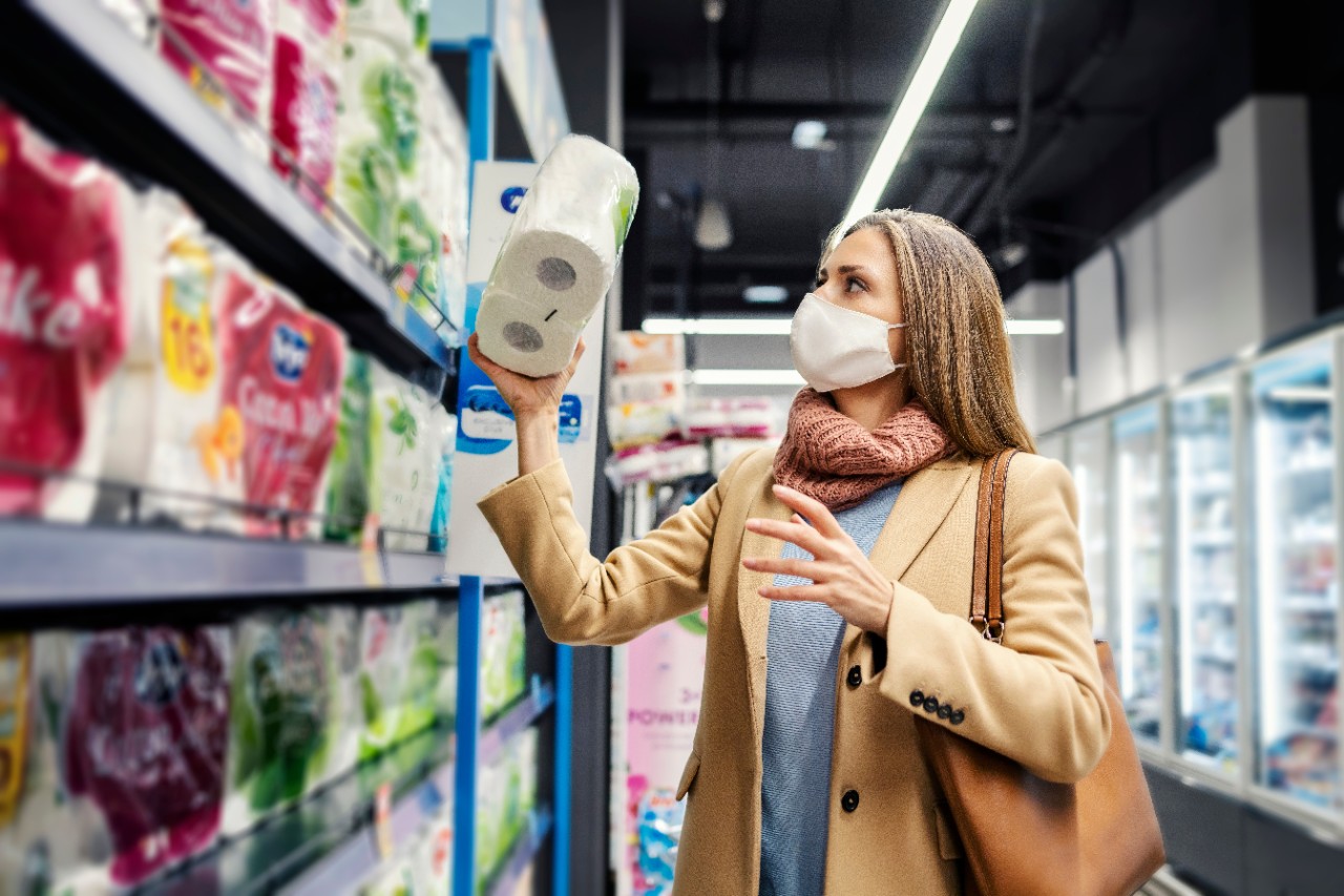 A woman in a mask and tan coat shops for groceries.