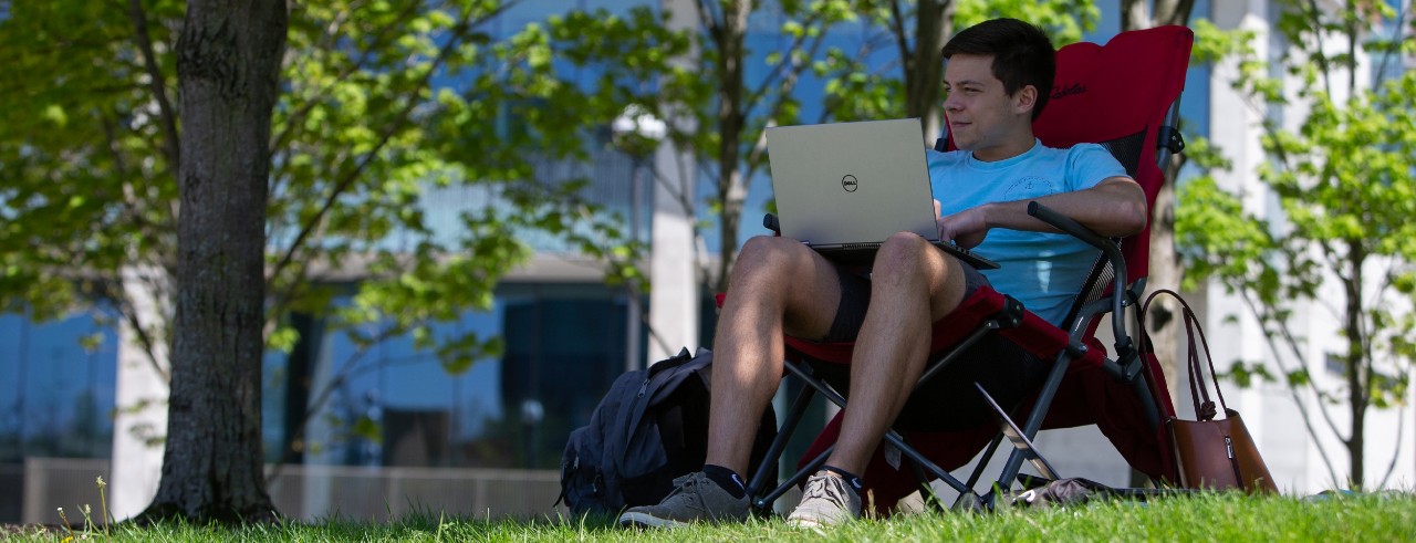 A student works on a laptop in the shade of a tree.