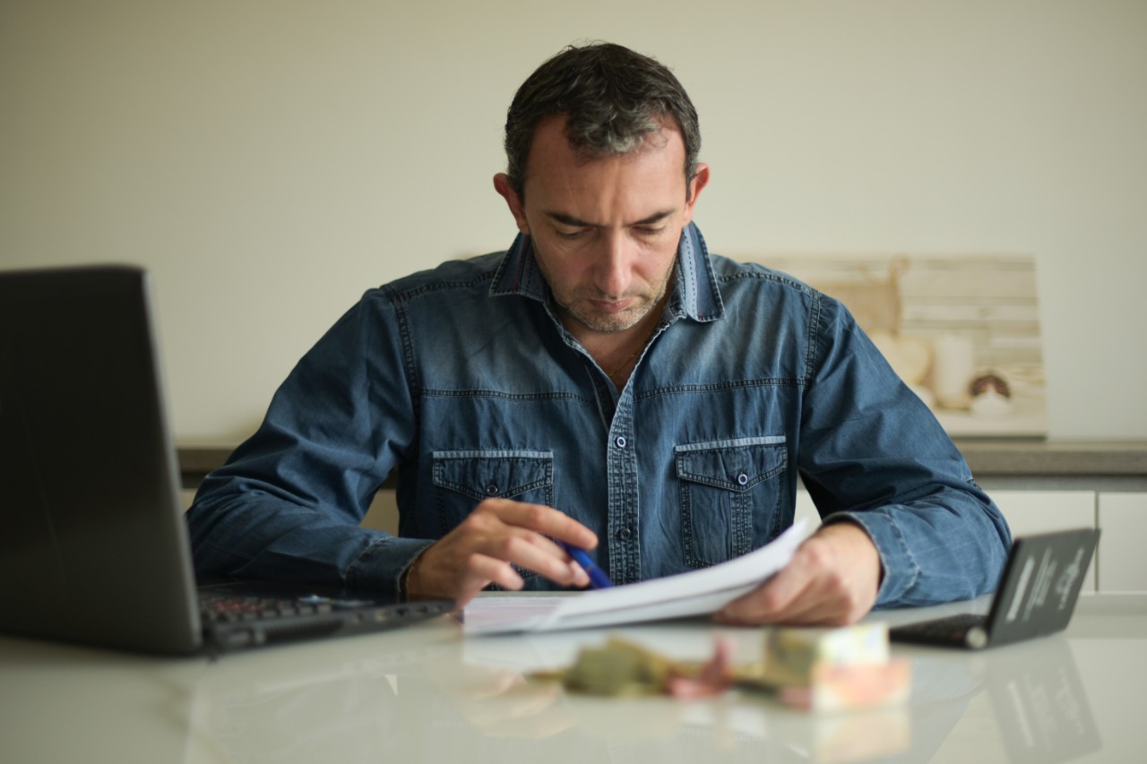 A man sits in front of a laptop in a blue shirt checking his bills.