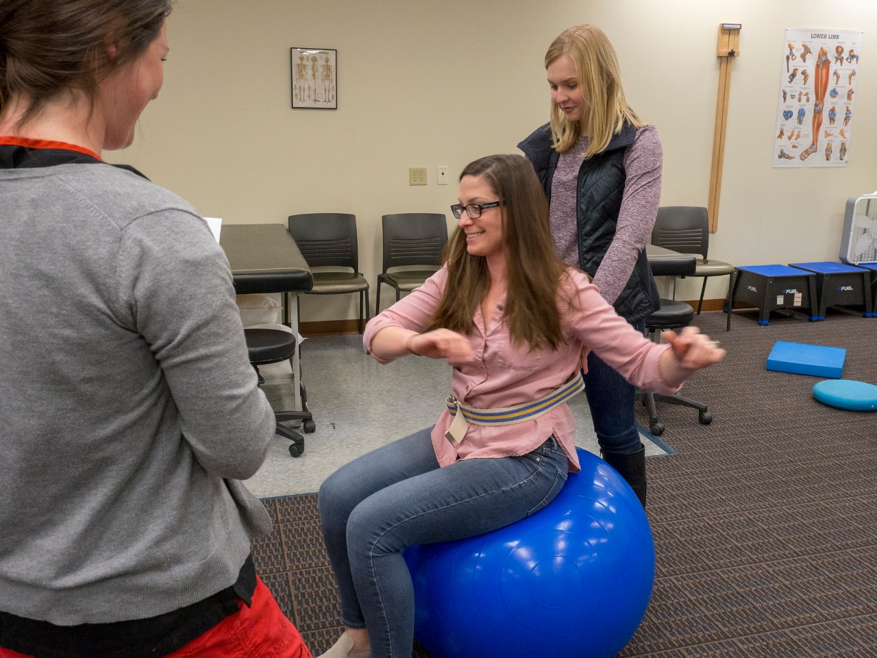 Two students learn physical therapy technique with a student seated on an exercise ball.