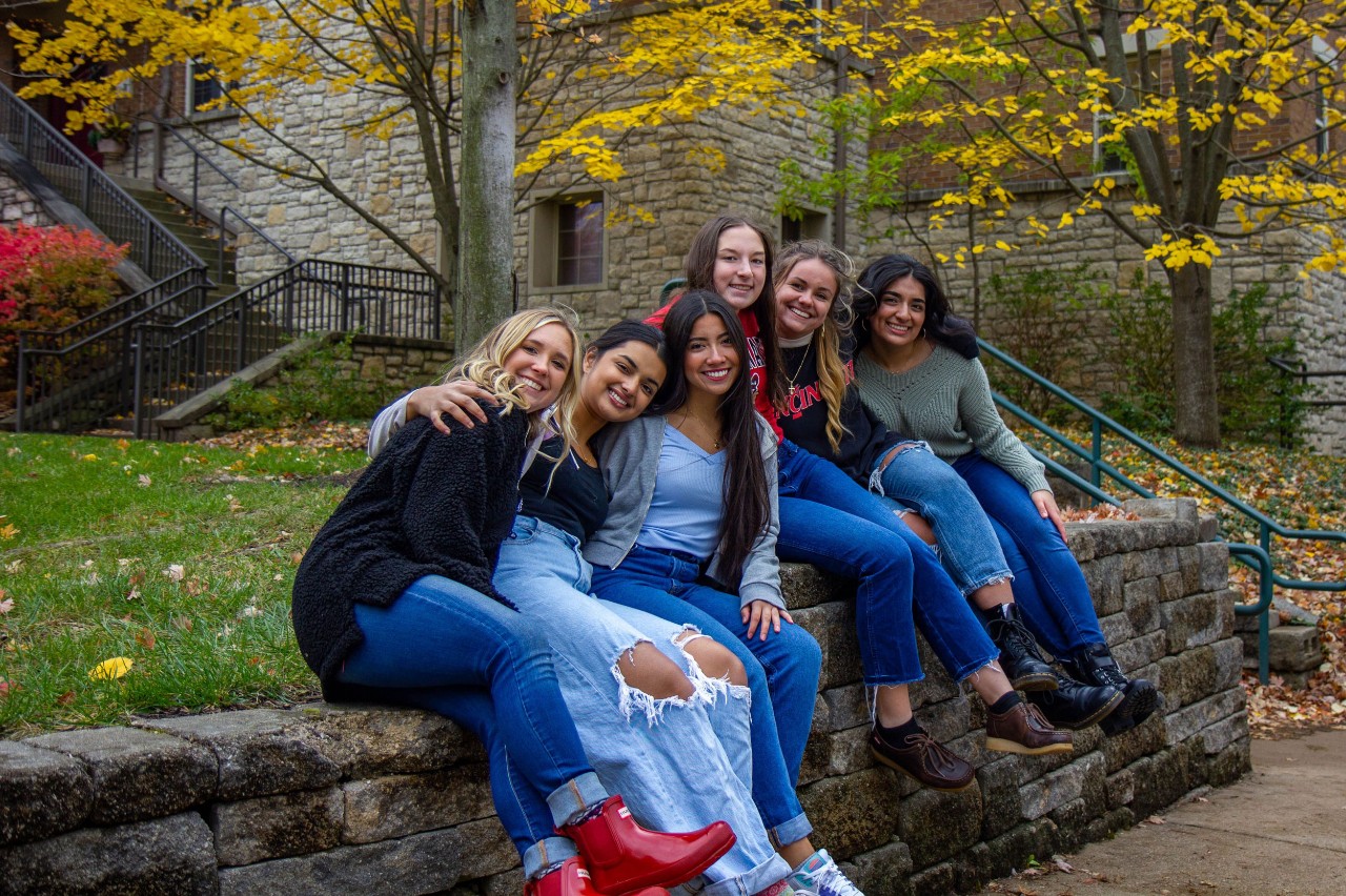 Six young women wearing jeans and long sleeve shirts gather and smile on a stone wall in front of a grassy lawn with a tree in the fall