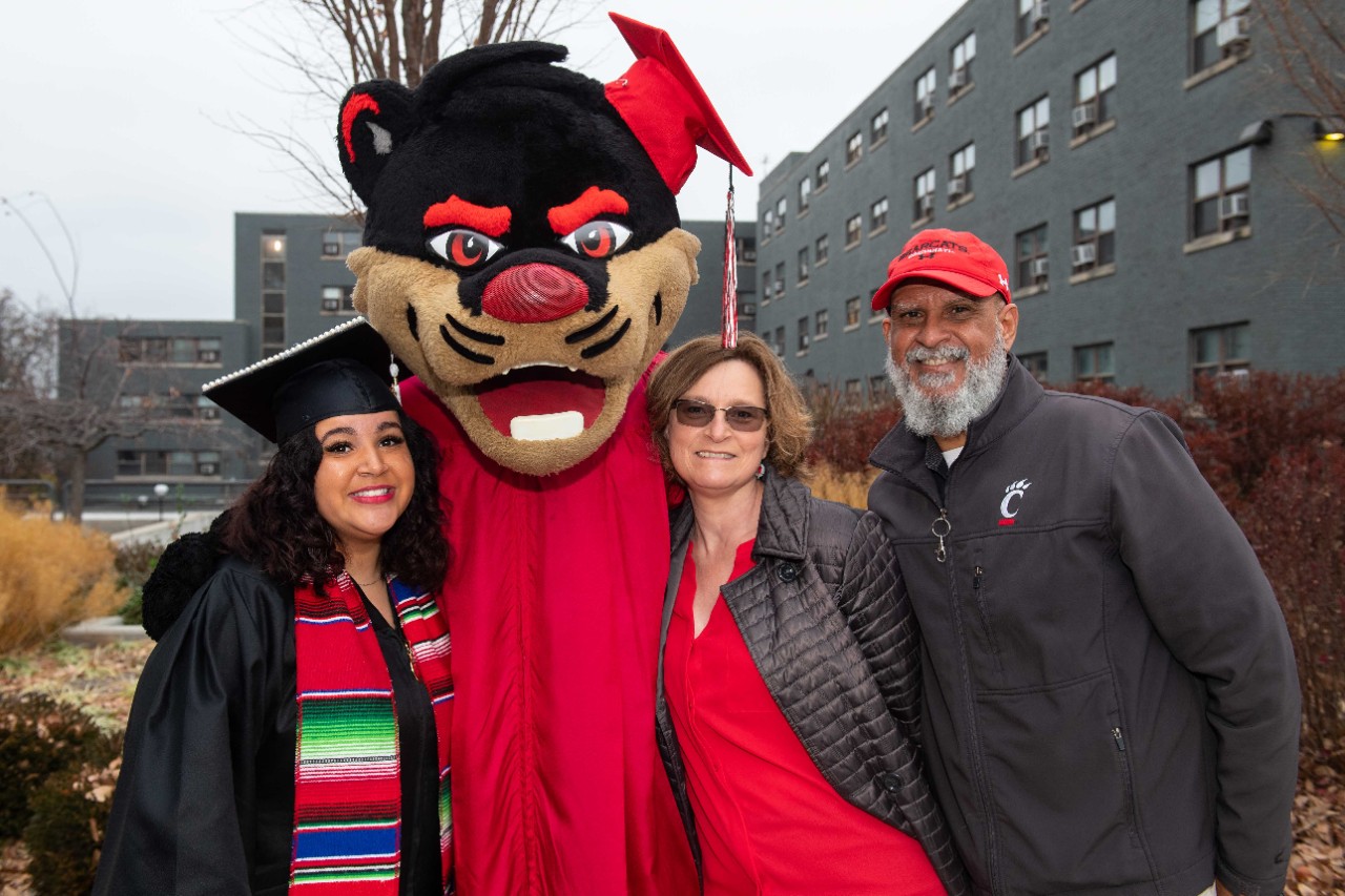A UC graduate in her cap and gown and her family takes a photo with the UC Bearcat, also wearing a mortarboard and gown, outside Fifth Third Arena.