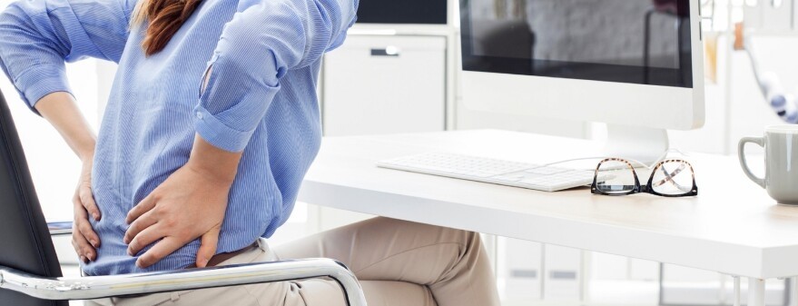 A woman seated at a desk holds her back in pain