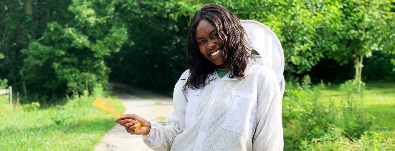 a woman in a beekeeper's protective equipment smiles at the camera