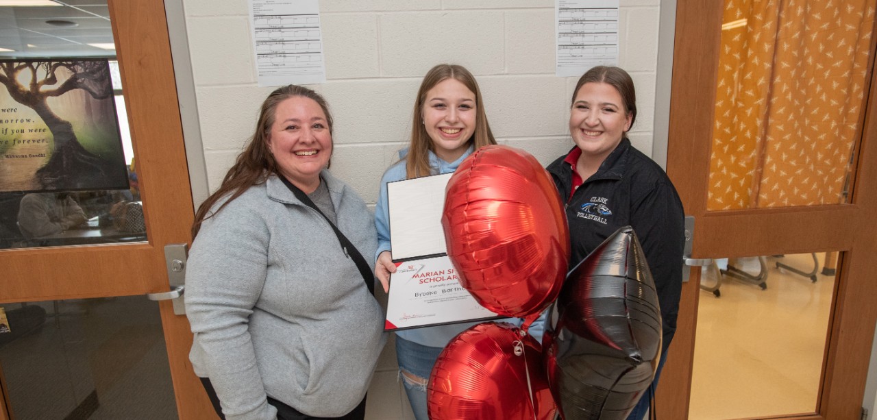 Brooke Bartholomew holds balloons and a certificate while celebrating her scholarship announcement with her family in a school hallway.