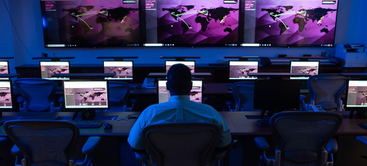 A person sits facing banks of computer screens and monitors on a wall with a map of the world.