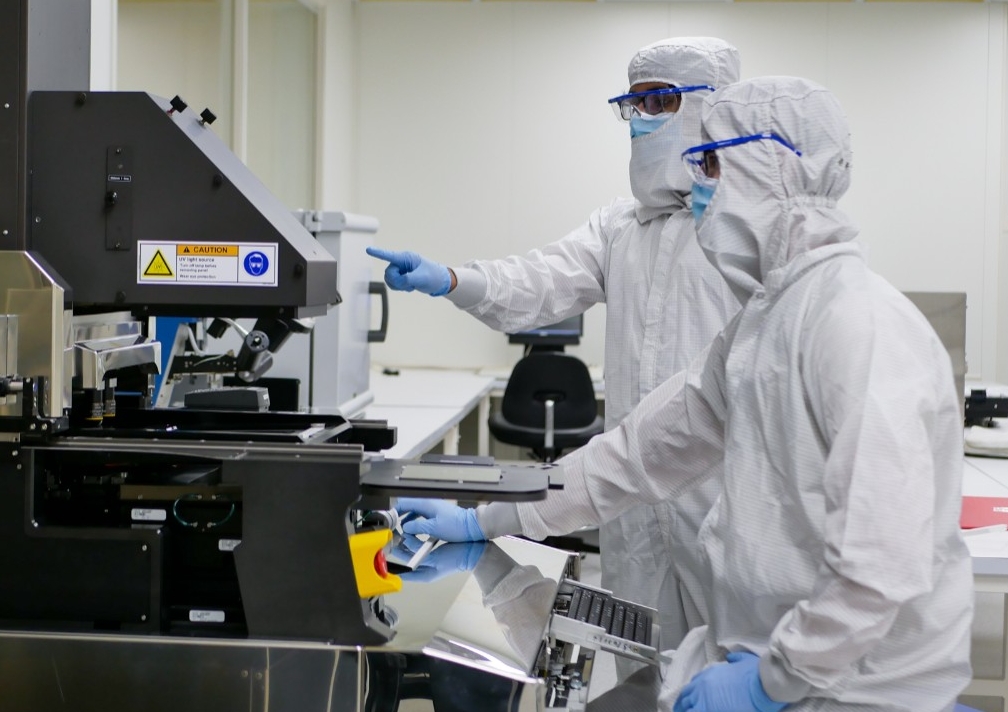 Two people wearing protective clothing from head to toe work with electronic equipment in a clean room.