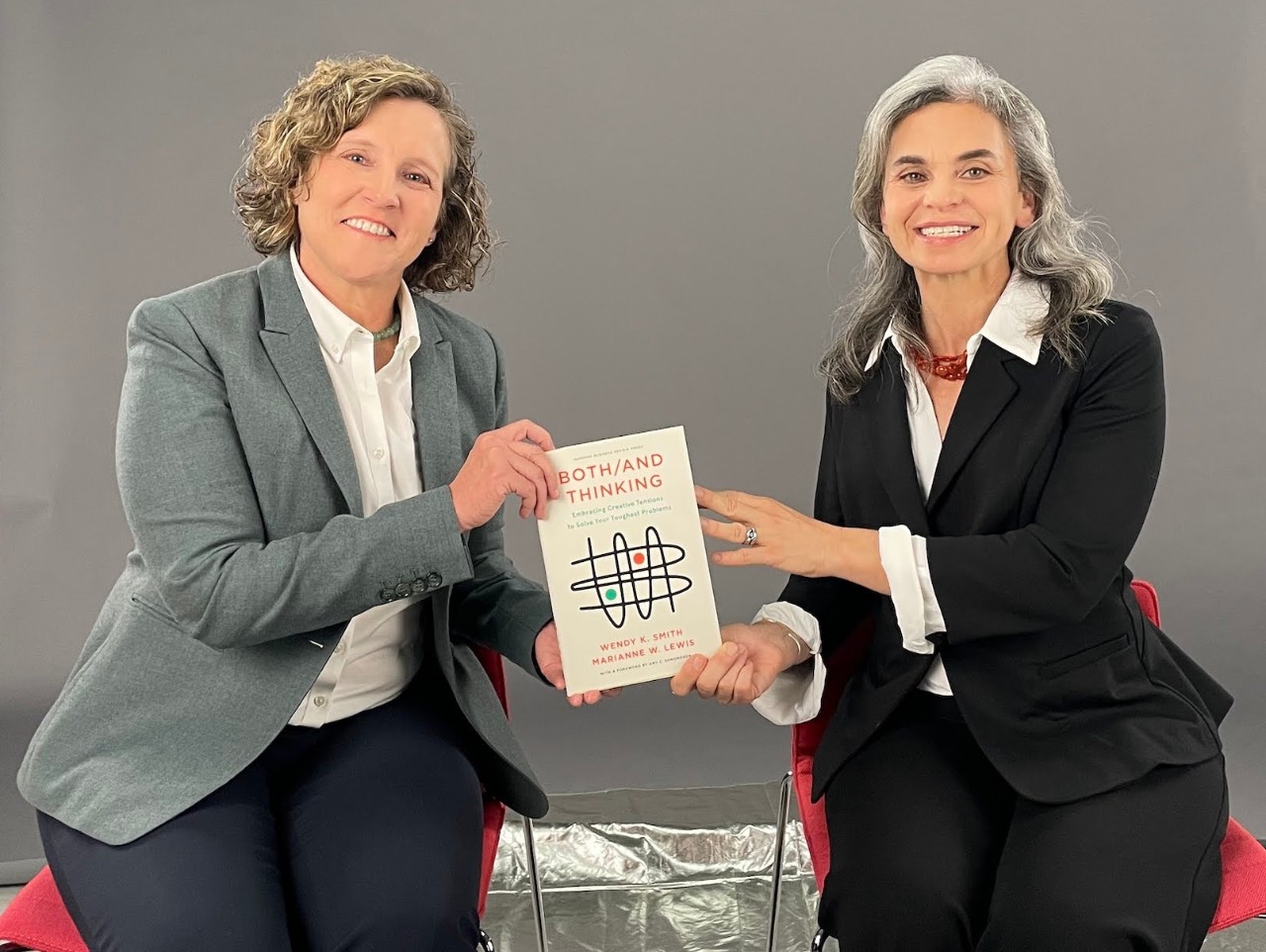 one woman with a white blouse and grey suit jacket and short blonde hair holds a book sitting next to a woman with long gray hair wearing a white blouse and black suit jacket in front of a gray background on a photo set