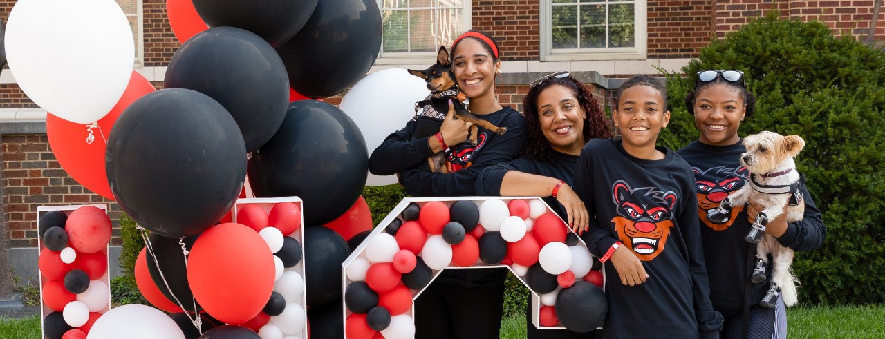 Image of a family at UC Family Weekend posed next to the UC display with balloons