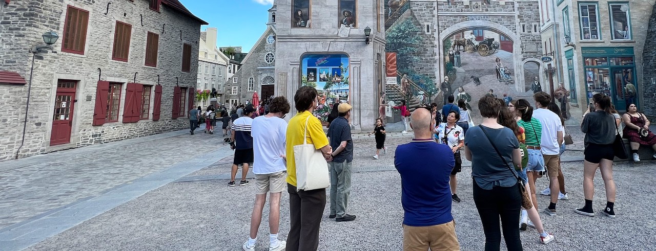 A group of people surrounds a tour guide in an old city square. The building behind the guide is painted with a trompe-l’oeuil mural of 18th-century French settlers and modern residents in windows and archways. To the left, people walk along a street of similar old buildings, completing the blend of old and new. 