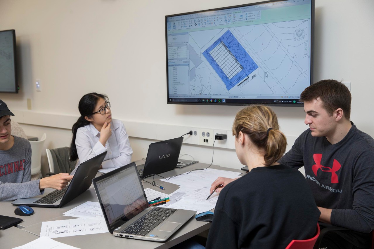 Three students work together on laptops while collaborating on a screen on a wall featuring blueprints.