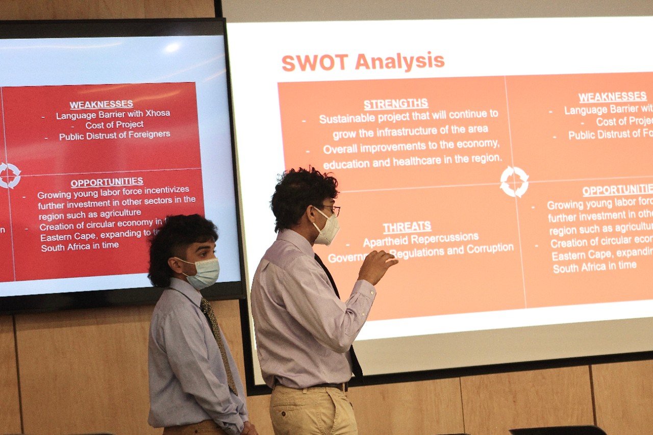Two UC students in face masks give a presentation in front of a projector image of lists with headings including strengths and weaknesses.