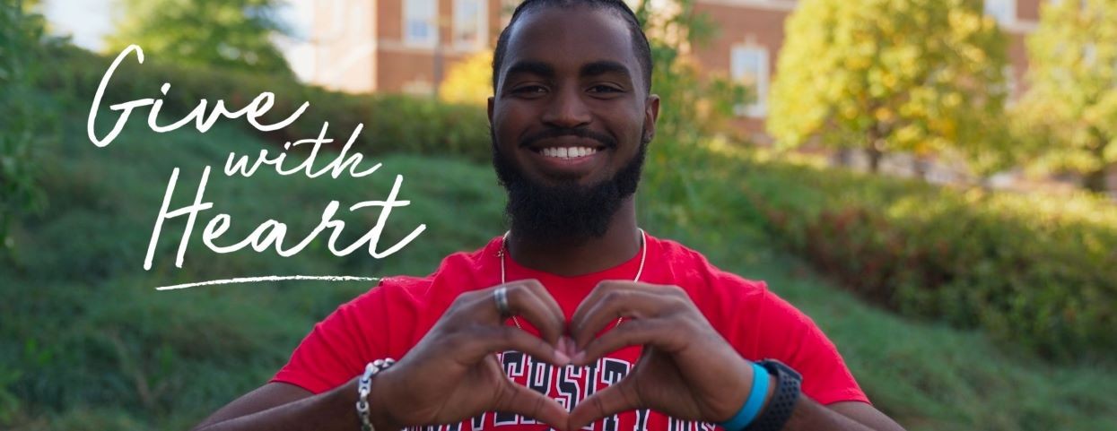 student smiling and making a heart with his hands with the words "give with heart"