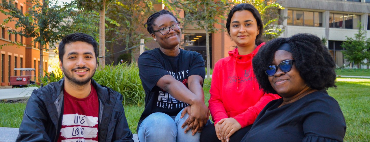 Four students wearing red and black t-shirts and sweatshirts, seated in a green space among campus building, smile toward the camera