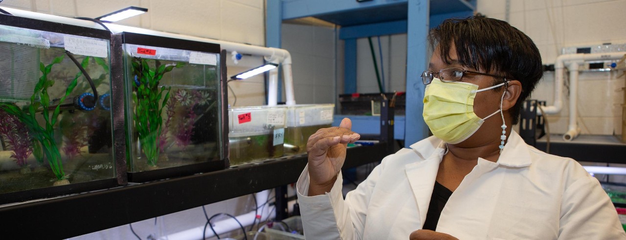Latonya Jackson stands in front of an aquarium of fish with a labcoat and face mask.