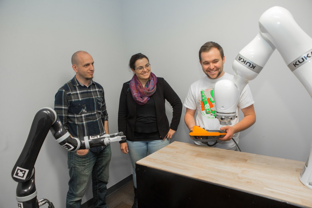 Three people stand around a robotic arm .