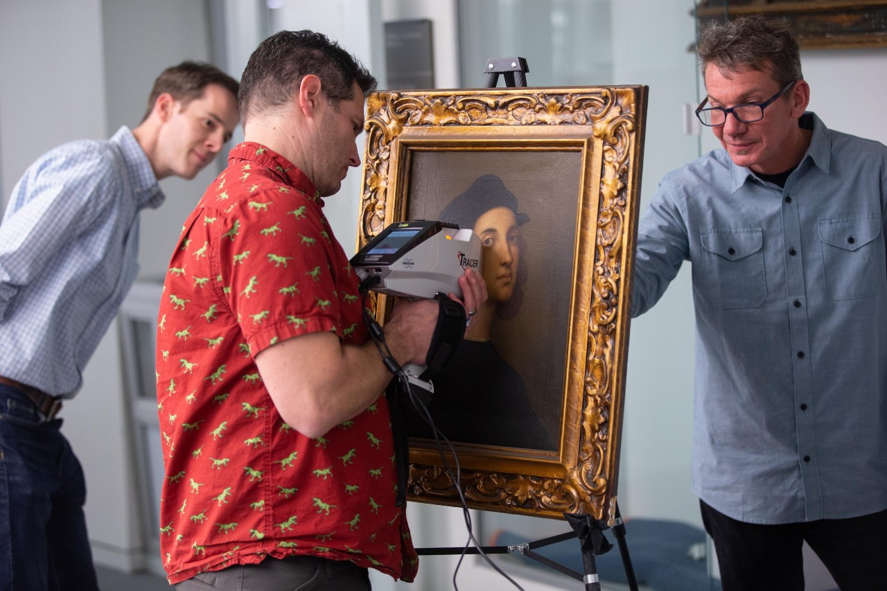 A geologist uses an XRF spectroscopy scanner to examine a painting while two people look on.