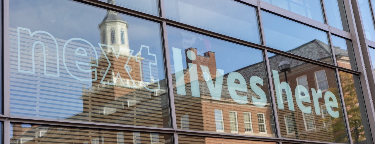 Window of University Pavilion shows Next Lives Here and reflection of campus.