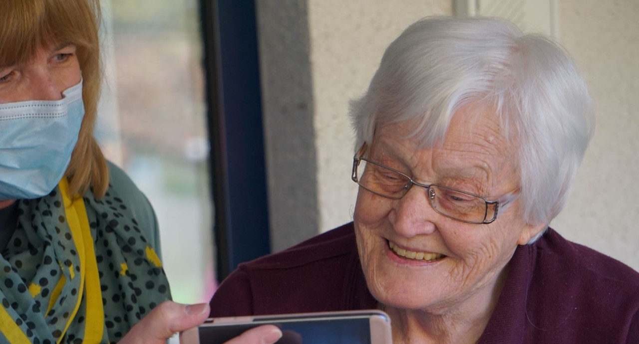 caregiver in mask showing and elderly woman something on a cell phone screen