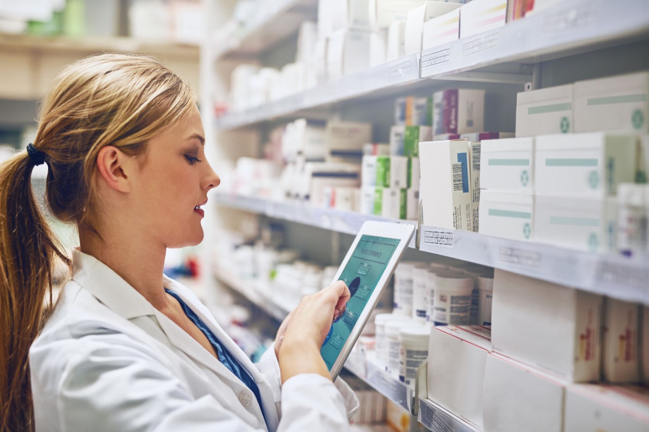 A female pharmacist looks at a tablet in front of shelves of medication