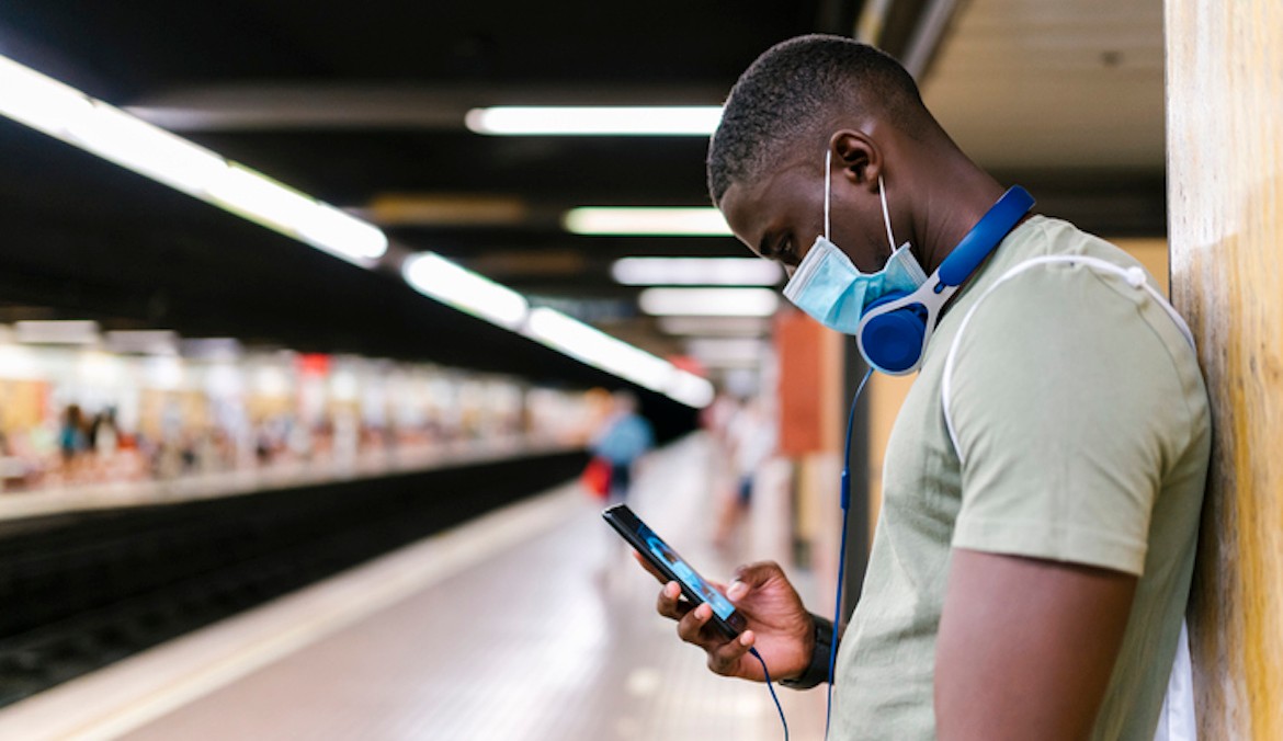 a man wearing a face mask checks his phone while standing at a train station