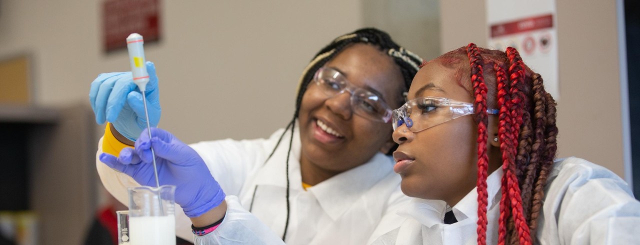 Two students wearing lab coats, goggles and gloves check the temperature of a mixture on a hot plate