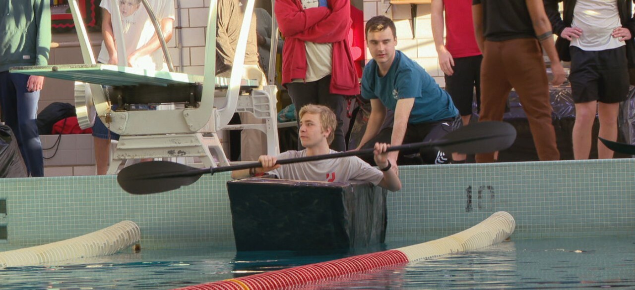 A student paddles a cardboard boat in a swimming pool.
