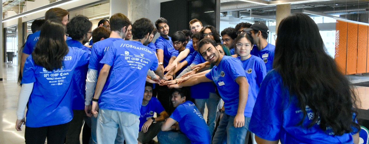 Several students wearing blue T-shirts stand together holding hands in a circle.
