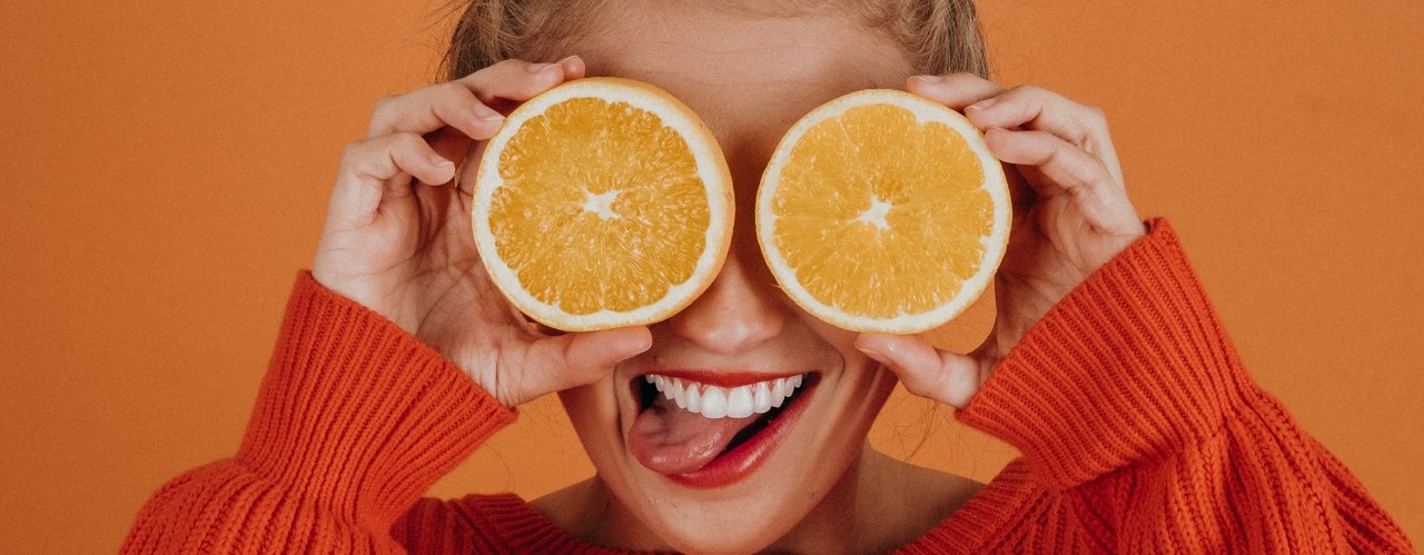 Woman stares forward with tongue sticking out holding two slices of oranges covering her eyes.