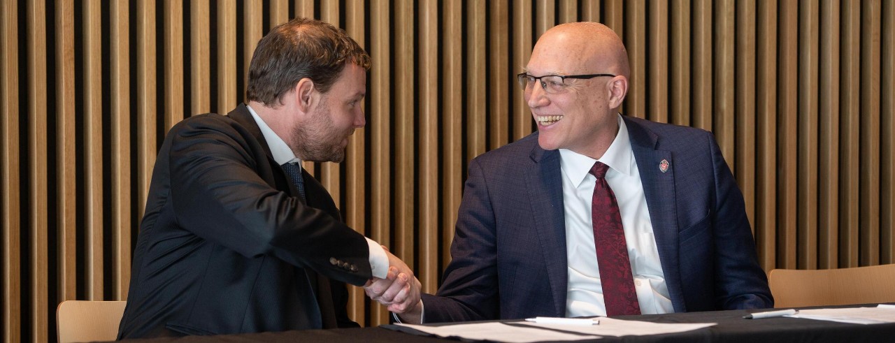 Two people in suits shake hands at a table in front of some documents.