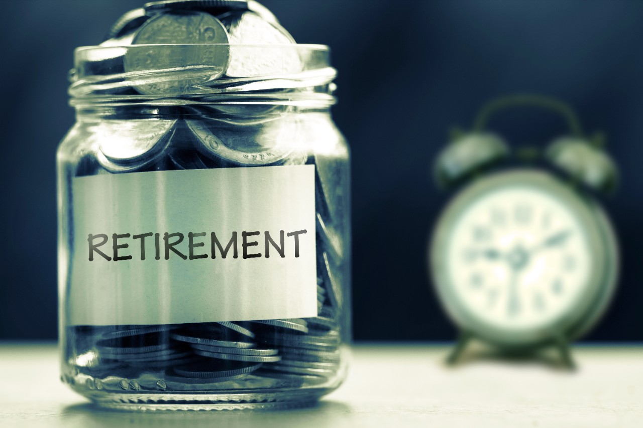 Close up coins in glass jar with sticker "Retirement" in foreground and alarm clock in background