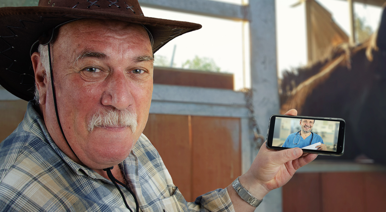 A cowboy in a barn holds up a cell phone with an image of a doctor in scrubs.