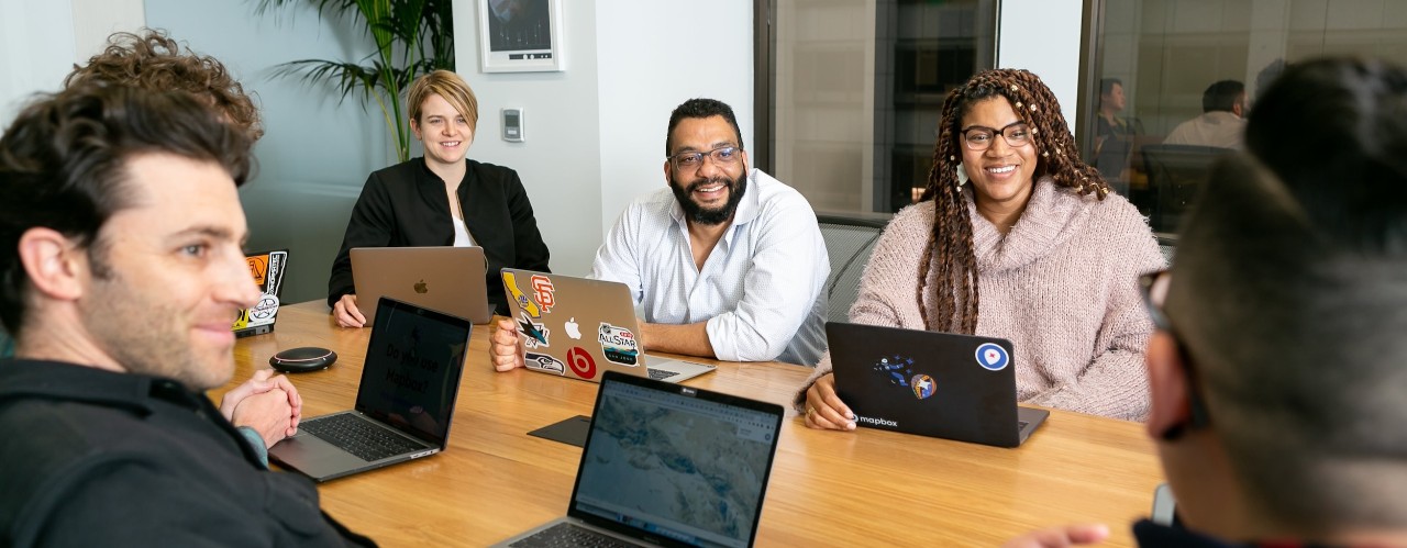 Men and women sit around a table with laptops.