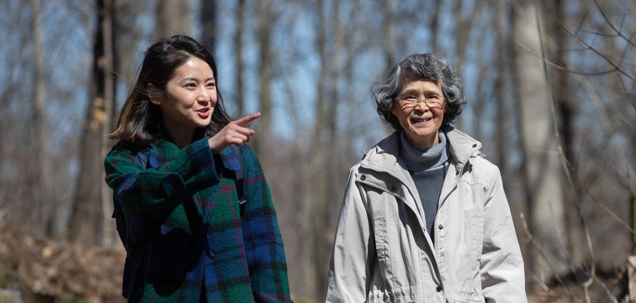 Two people in coats walk through the woods while talking.