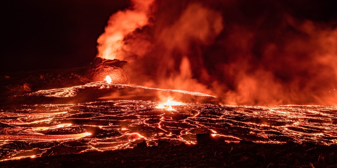 A volcano erupts at night creating a glowing pool of lava covered in plumes of sulfur.