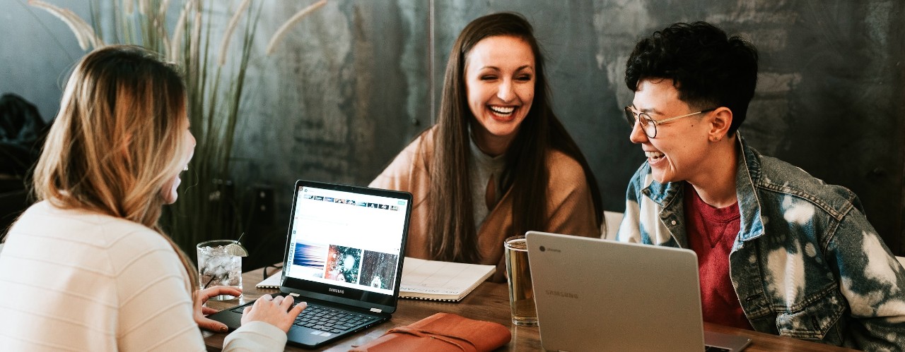 Three people sit at a table smiling over their laptops.