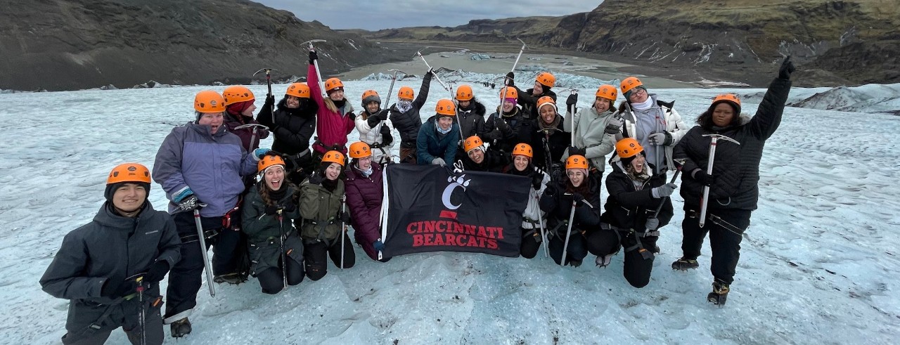 Students from the Travel Writing in Iceland course raise ice picks in the air and hold a UC flag while hiking on a glacier.