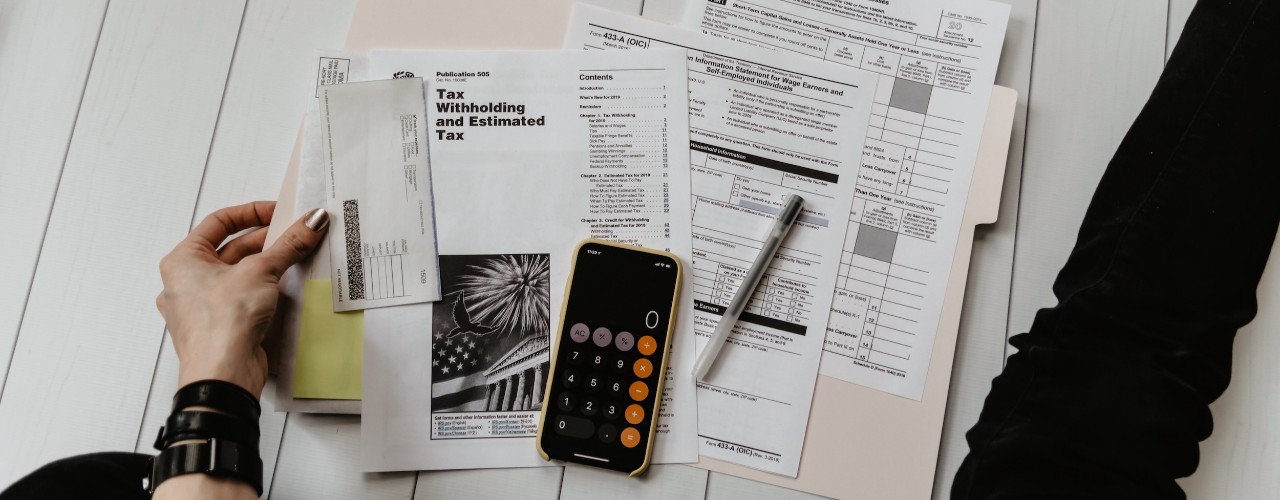 Close-up of IRS tax documents on a floor with calculator, pen and a person's hand and leg on either side of the documents.