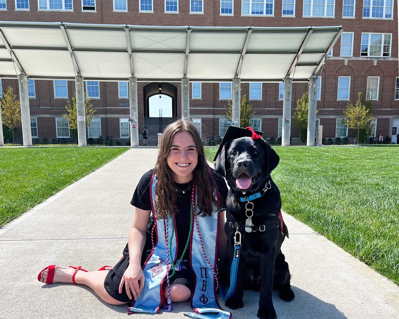 UC College of Nursing graduate Sara van de Sandt wearing a Commencement stole with black Labrador Retriever Lumen wearing a cap