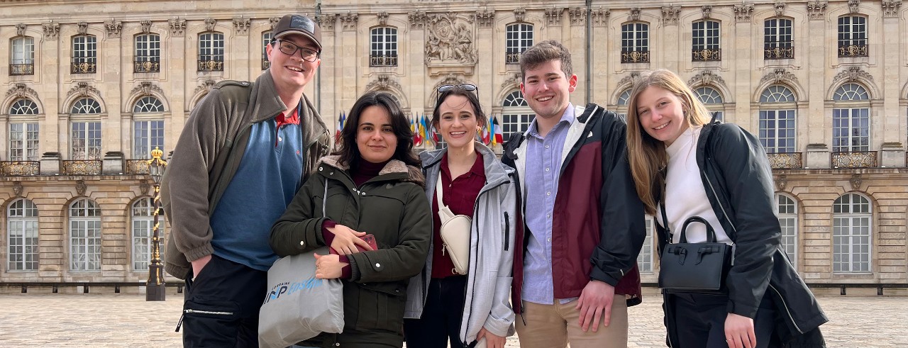 5 students pose smiling in the town square of Nancy, France, in front of a light-colored stone building with many arched windows and traditional decorative architectural elements