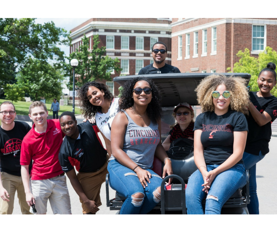 Students gathered on UC MainStreet sitting on the back of a car. Old Chem and Swift Hall are behind them.