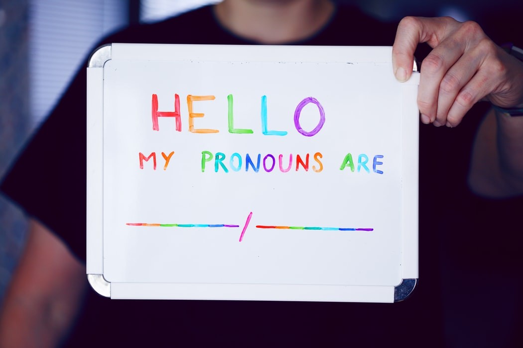 A person holds up a dry erase sign that says "Hello, my pronouns are __/__" written in rainbow lettering