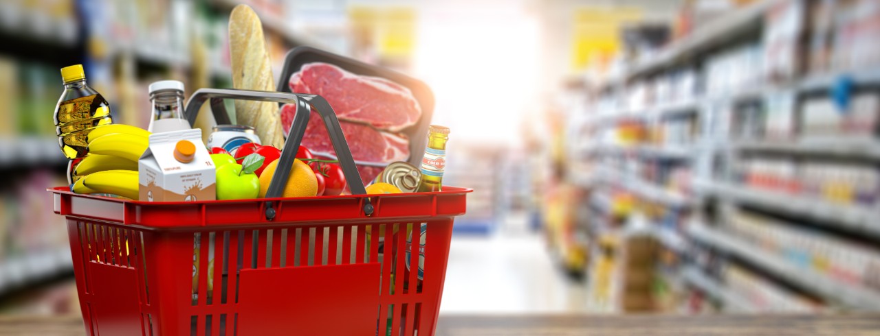 Red grocery basket filled with groceries sits on a table inside of a grocery store.