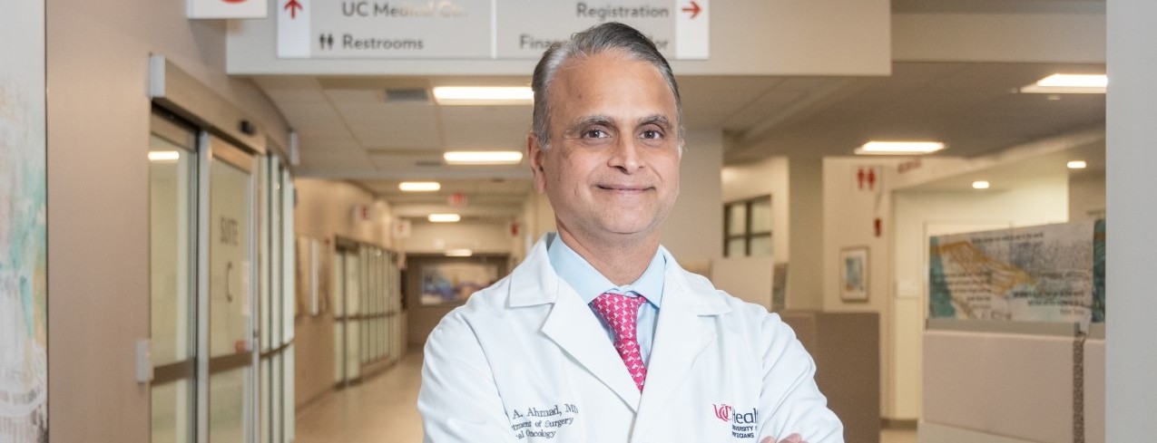 Dr. Ahmad stands and smiles wearing a lab coat with his arms crossed inside the Barrett Cancer Building