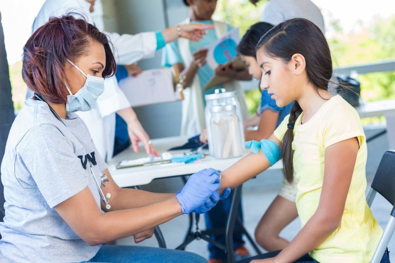 Public health nurse in a mask administering a vaccine to a young girl 